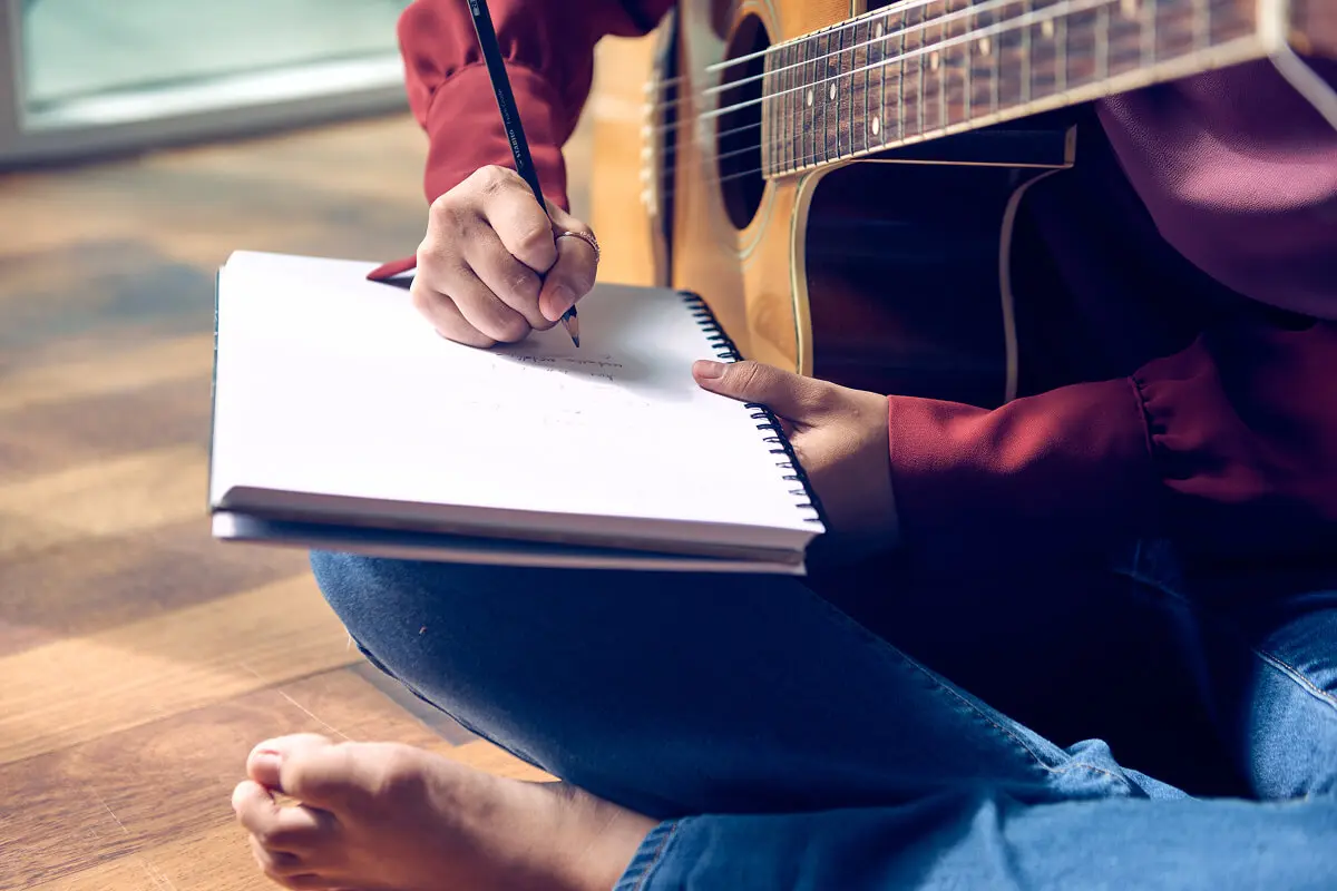 Person writing music with guitar on lap.